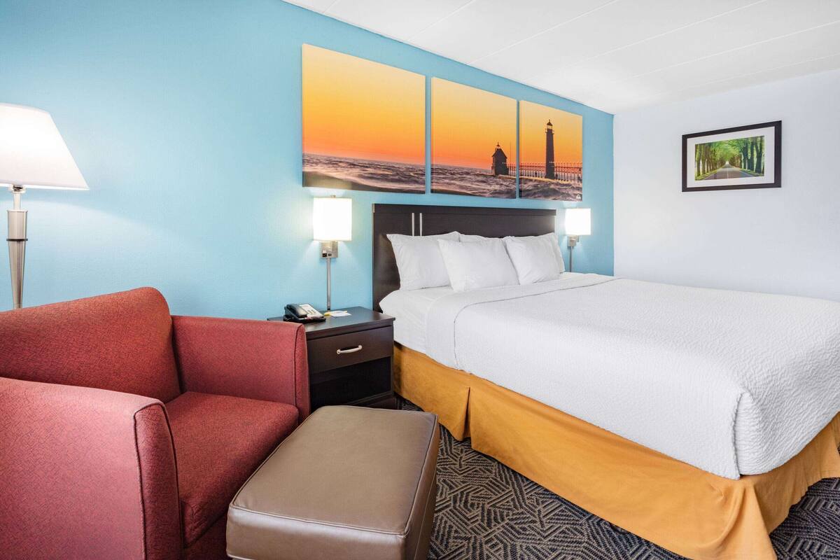 A hotel room with a bed, a red armchair, a nightstand with a lamp, and a painting of a lighthouse on the wall.