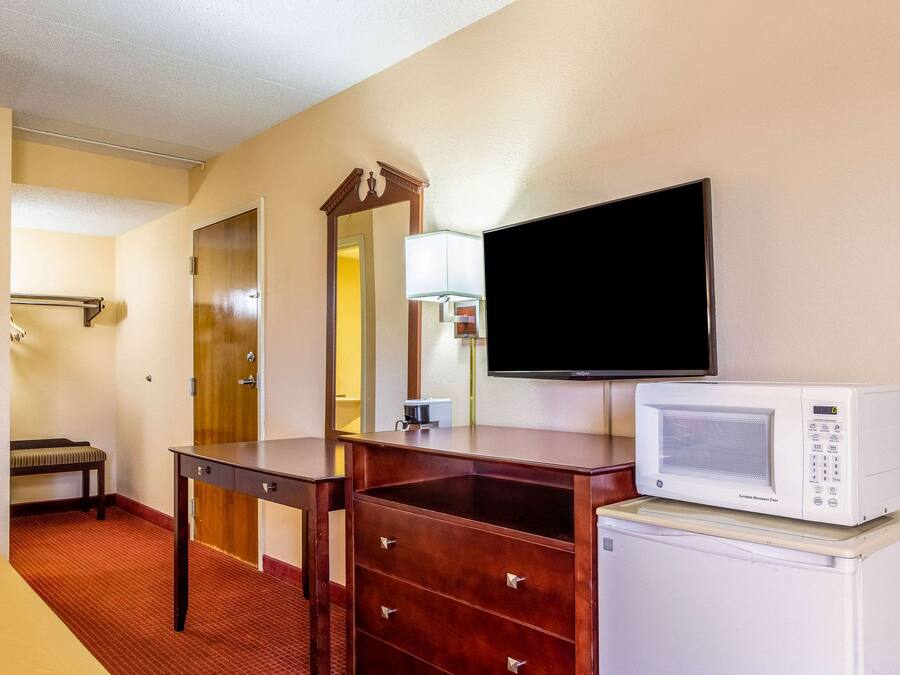 A hotel room with a wooden dresser, a microwave, a television, a bench, and a door.