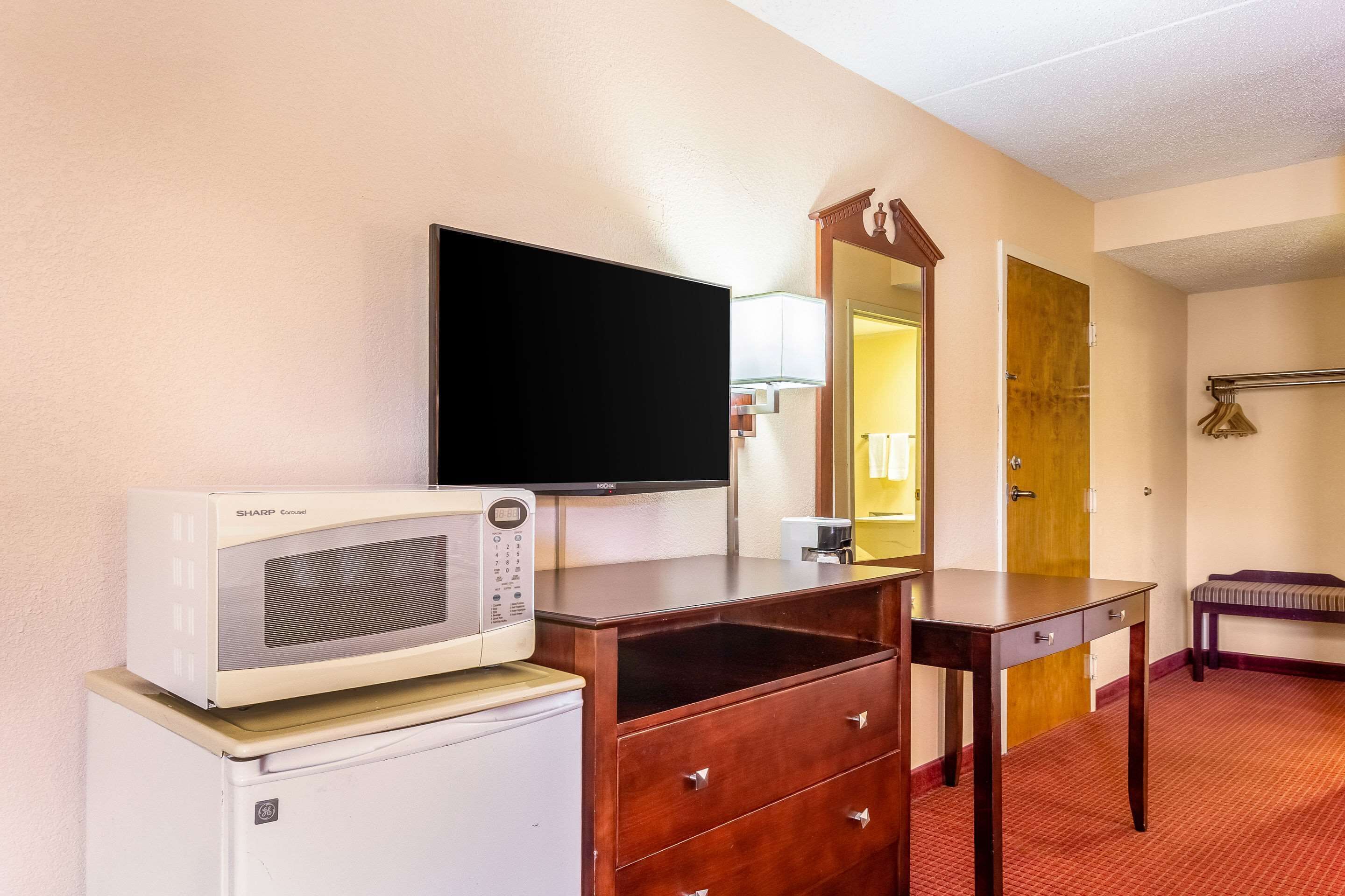 A hotel room with a microwave, a television, a wooden dresser, a mirror, a bench, and a door.