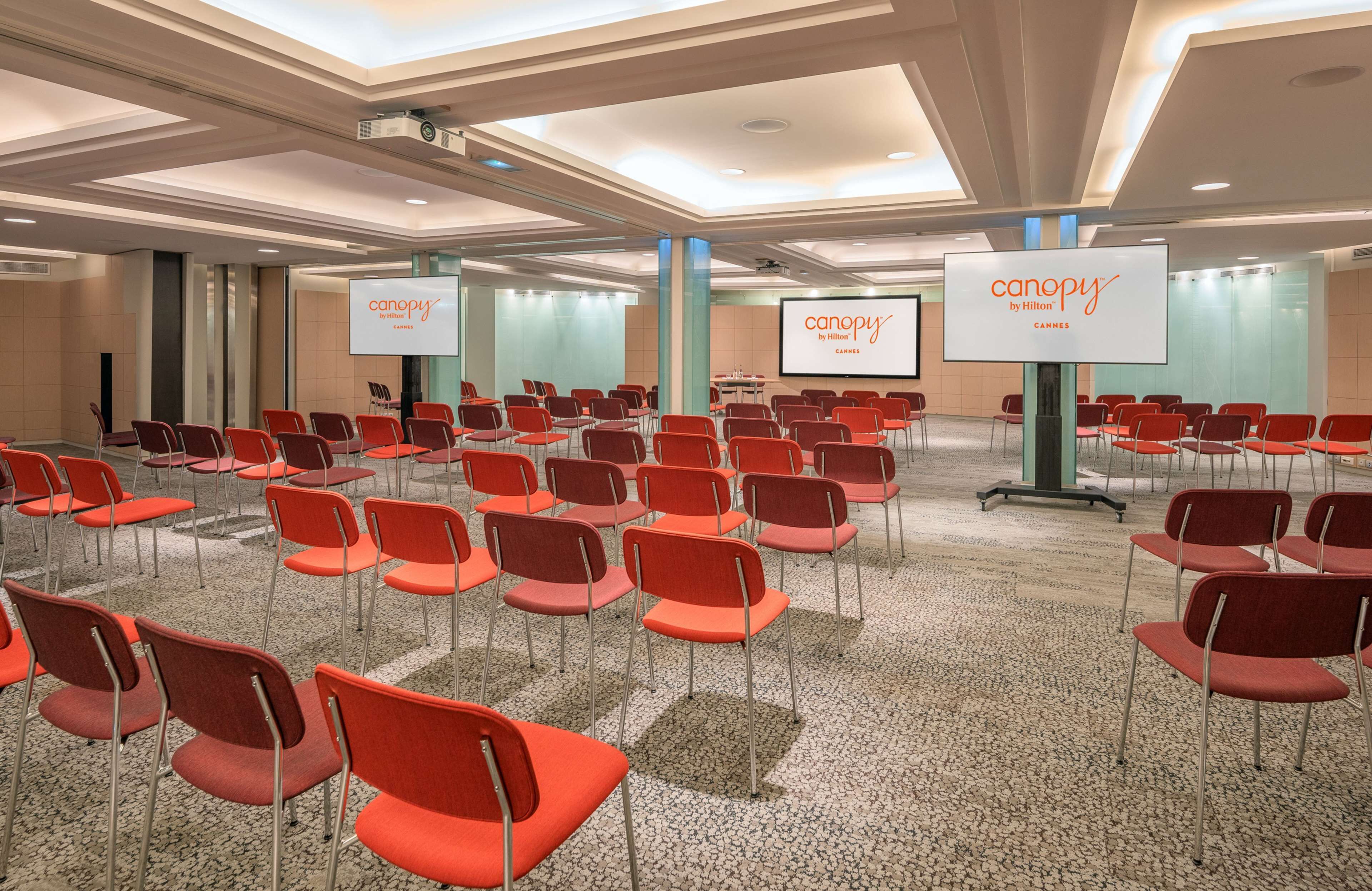 A conference room with rows of red chairs, a large screen displaying 'canopy by Hilton Cannes', and a modern ceiling design.