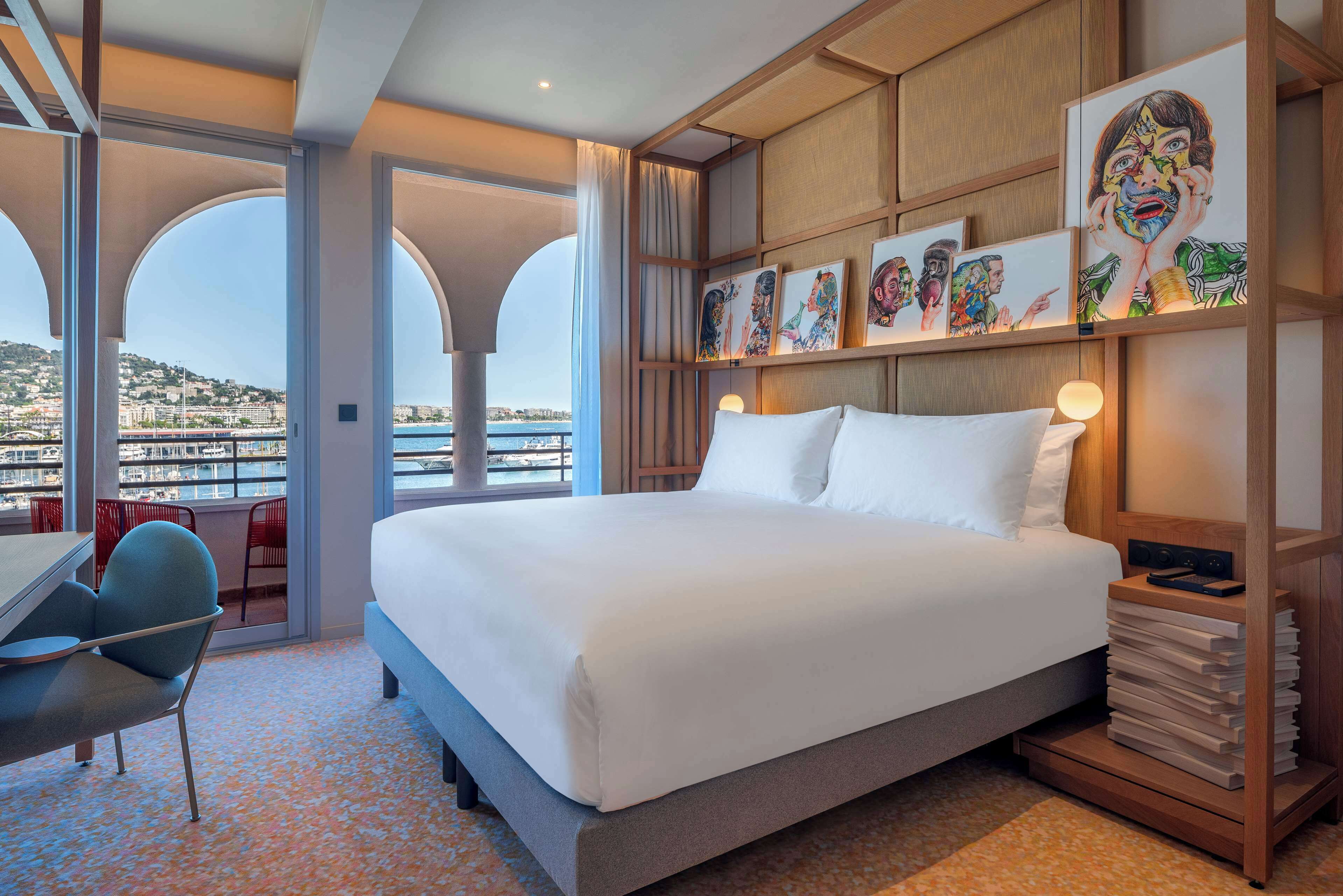 A hotel room with a large bed, a desk, a chair, and a view of a cityscape through large windows.