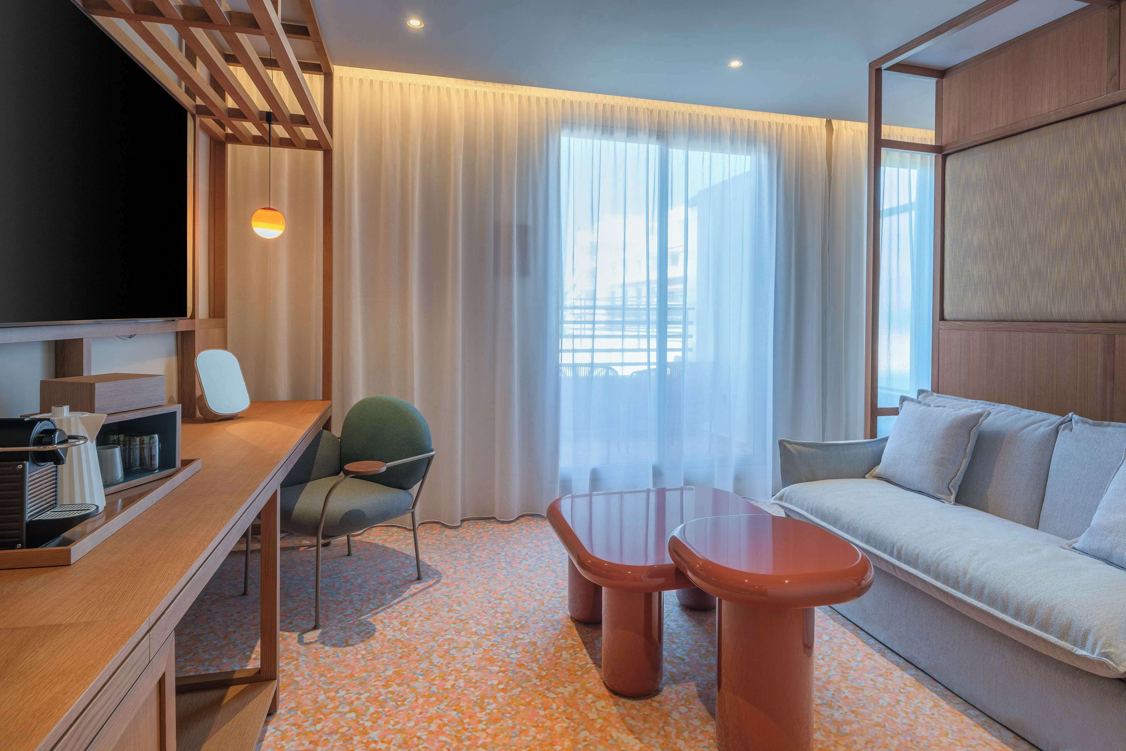 A modern hotel room with a wooden desk, a comfortable sofa, a television, and a large window with sheer curtains.