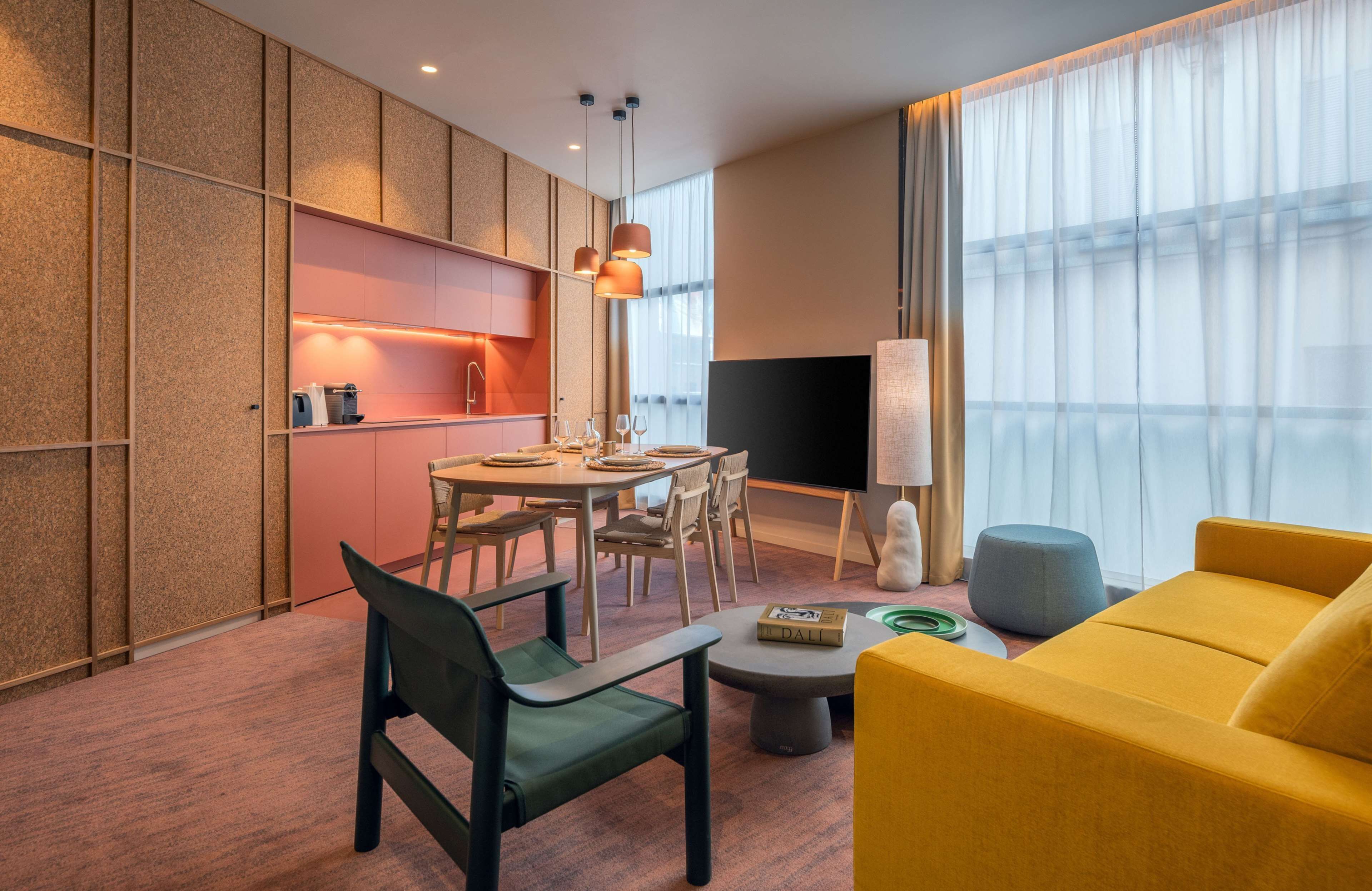 A modern living room with a yellow sofa, a dining table with chairs, a TV, and a kitchen area with a sink and cabinets.