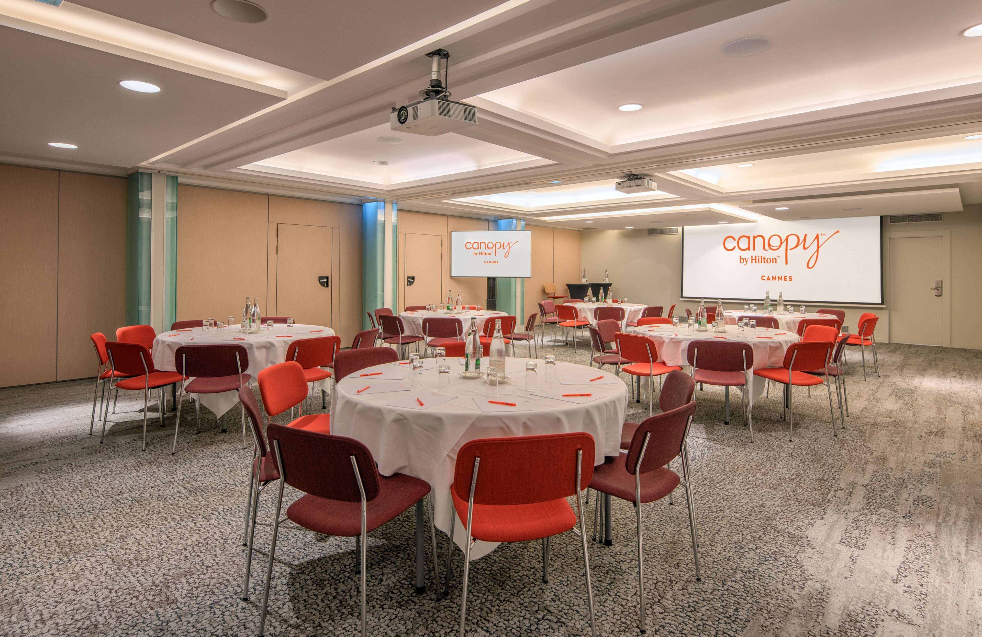 A conference room with multiple round tables, red chairs, and a large screen displaying the hotel's name.