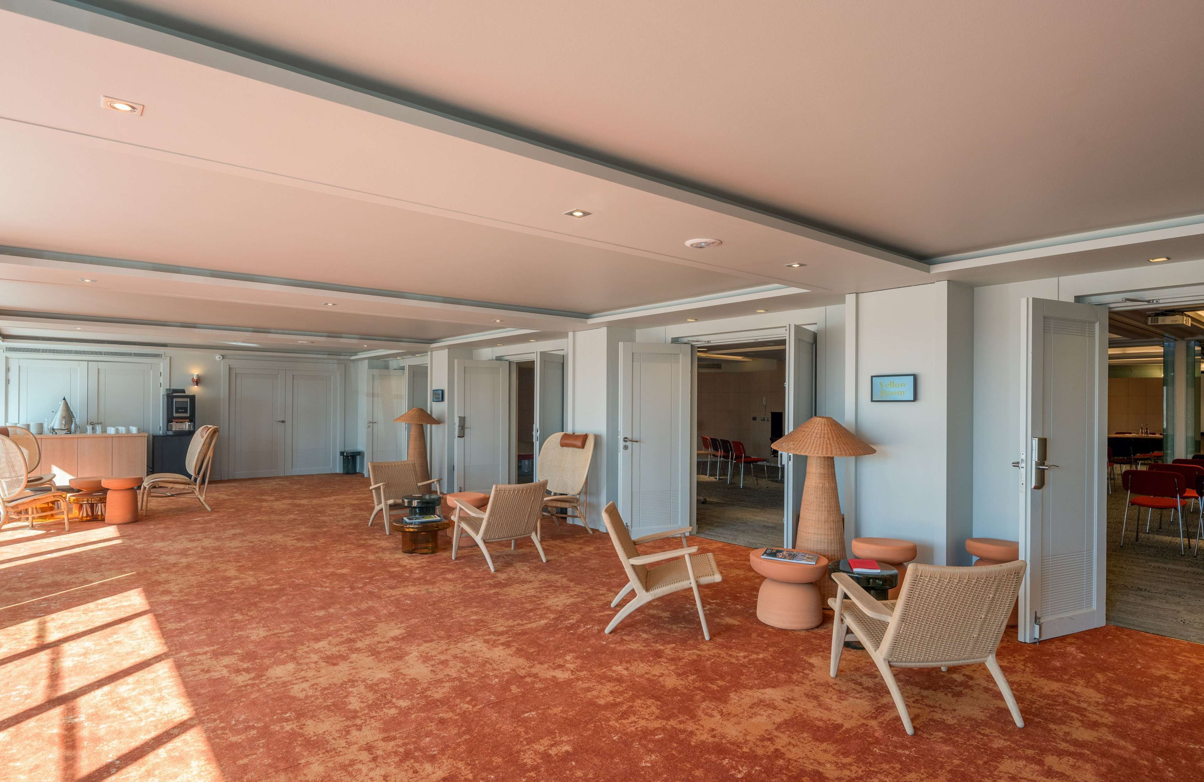 A spacious room with a red carpet, several wicker chairs, a table, and a lamp.