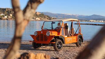 An orange vehicle with "canopy by Hilton" branding parked on a sandy beach with a body of water and mountains in the background.