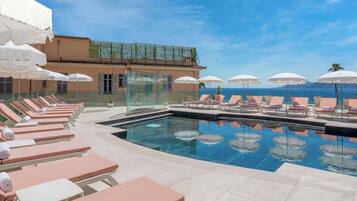 A poolside area with lounge chairs and umbrellas, a clear view of the sea, and a building with a tiled roof.