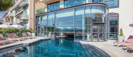 A modern hotel with a glass-enclosed pool area, sun loungers, and a curved glass facade.