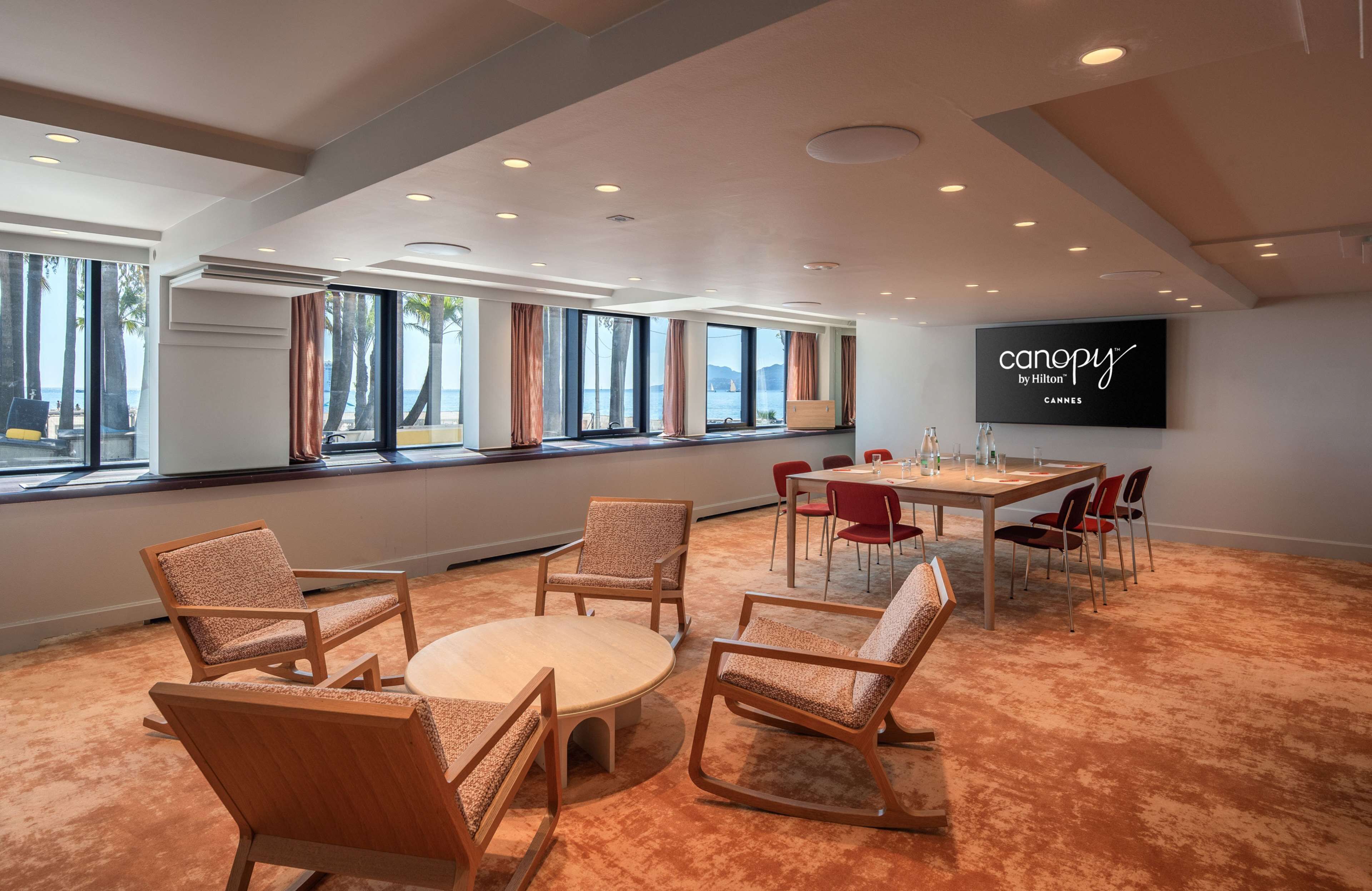 A conference room with a large screen displaying "canopy by Hilton" and "Cannes". The room has large windows with curtains, a round table with chairs, and a carpeted floor.