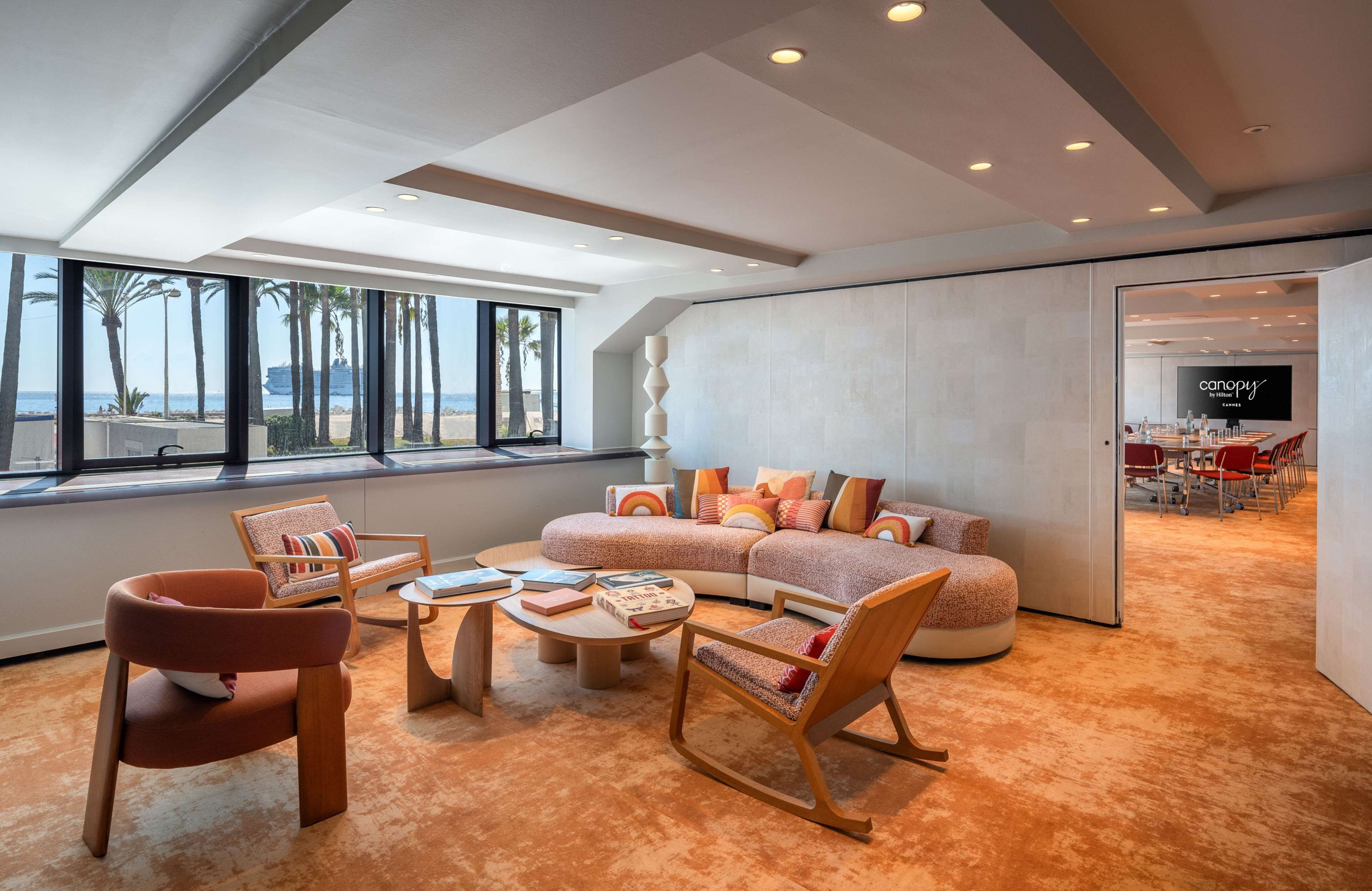 A modern living room with a large window, a sectional sofa, a coffee table, and a dining area visible through an open door.