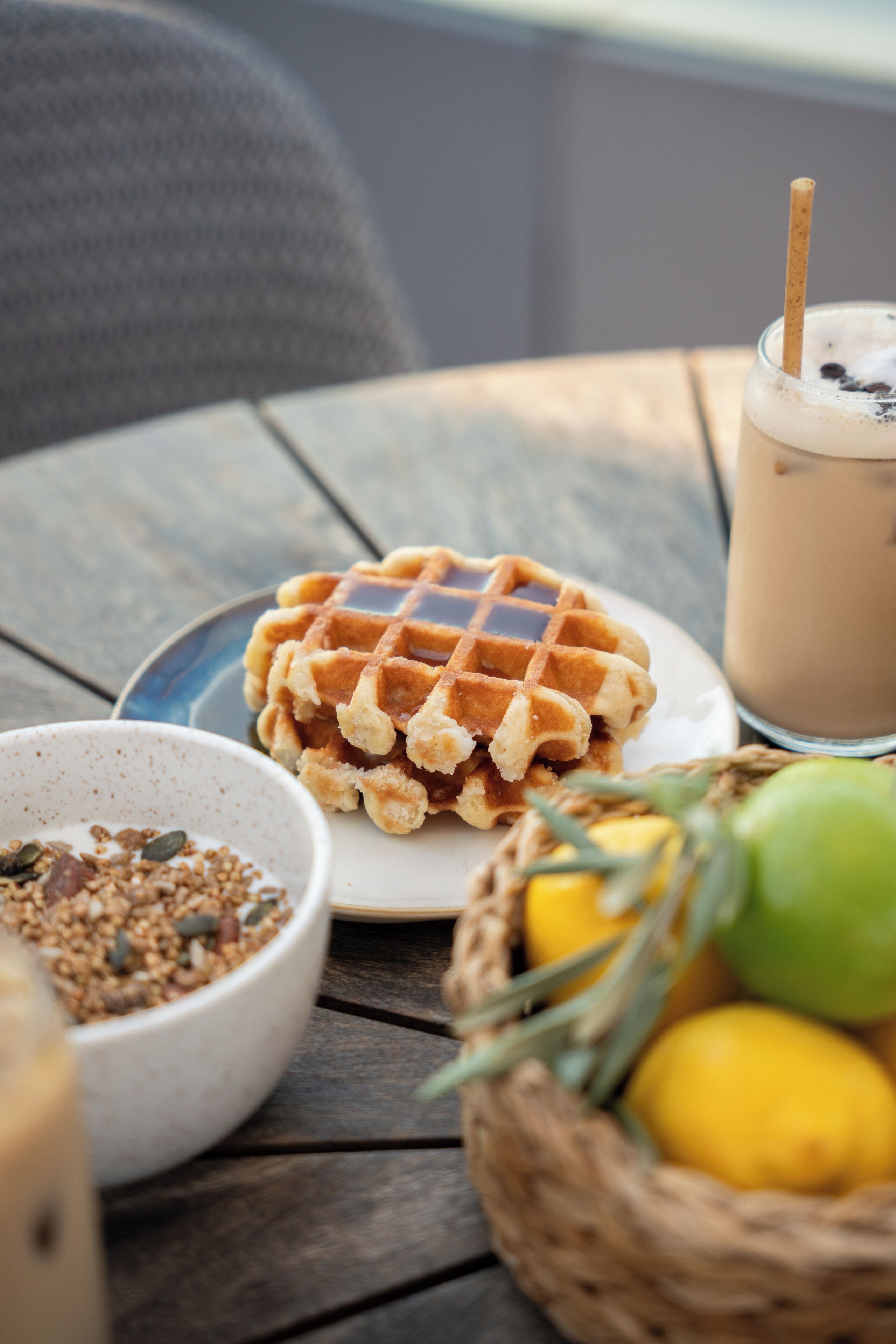 Ein Waffel auf einem Teller, ein Glas Eistee, eine Schüssel mit Granola und ein Korb mit Zitronen und Limetten auf einem Holztisch.