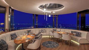 A modern dining area with a round table and chairs, a sofa, and a view of the city at night. - Canopy by Hilton Cannes (Cannes)