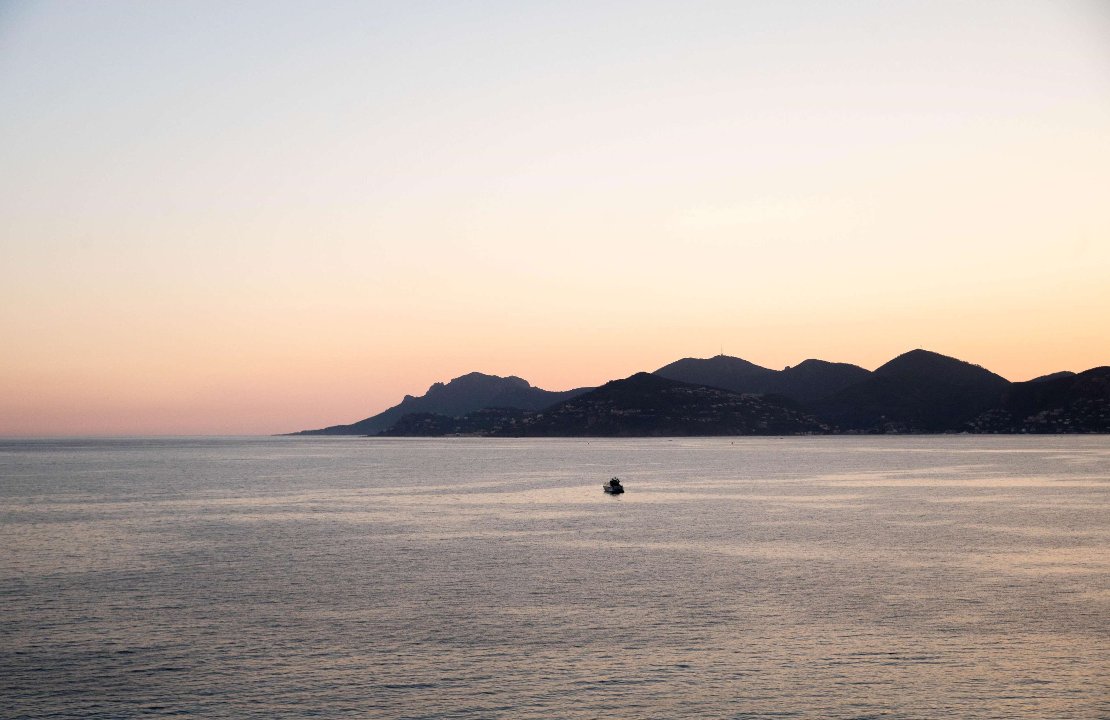 Une mer calme, avec un bateau à l’horizon et des montagnes en arrière-plan.