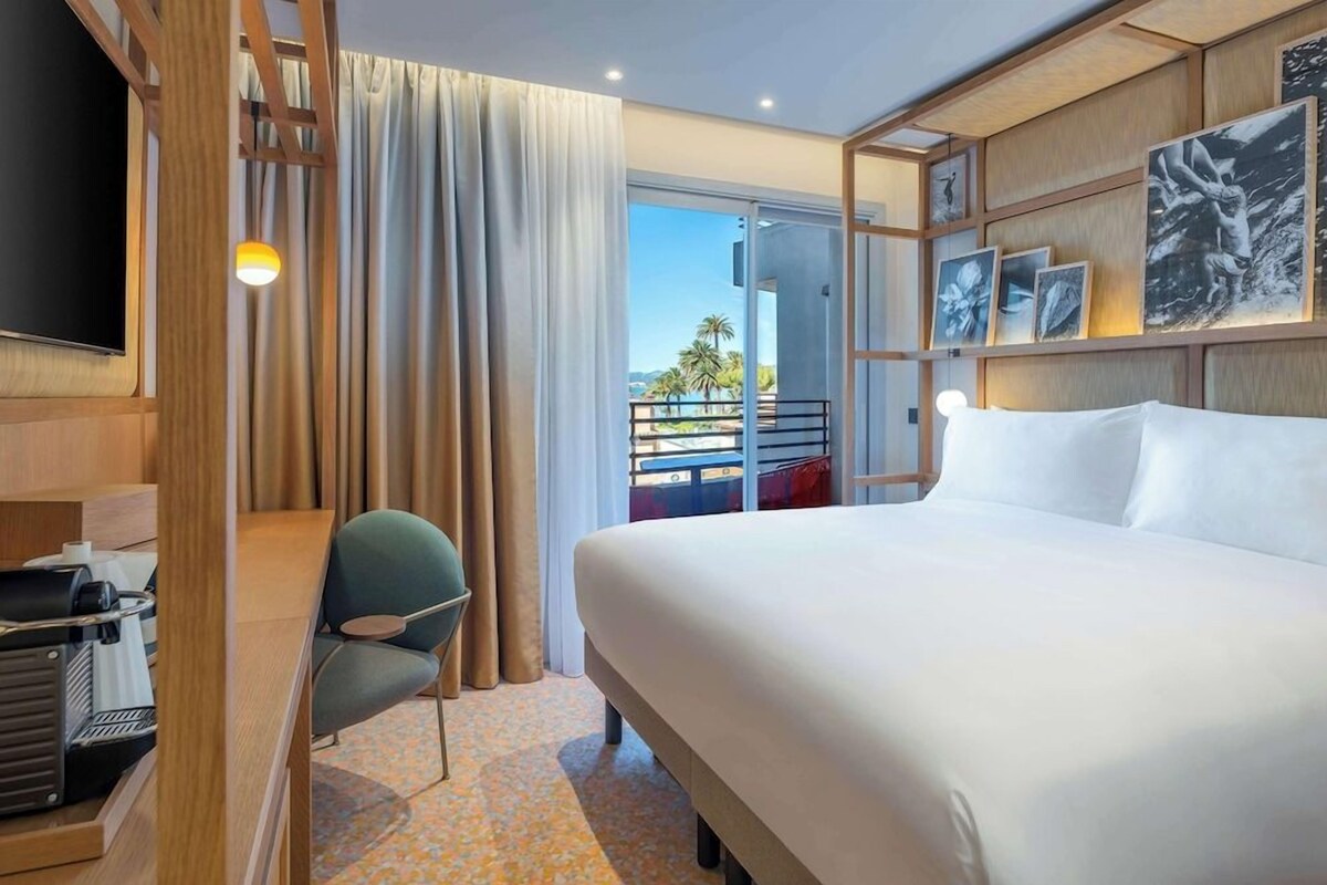 A hotel room with a bed, a desk with a coffee machine, a chair, a balcony with a view of palm trees, and artwork on the wall.