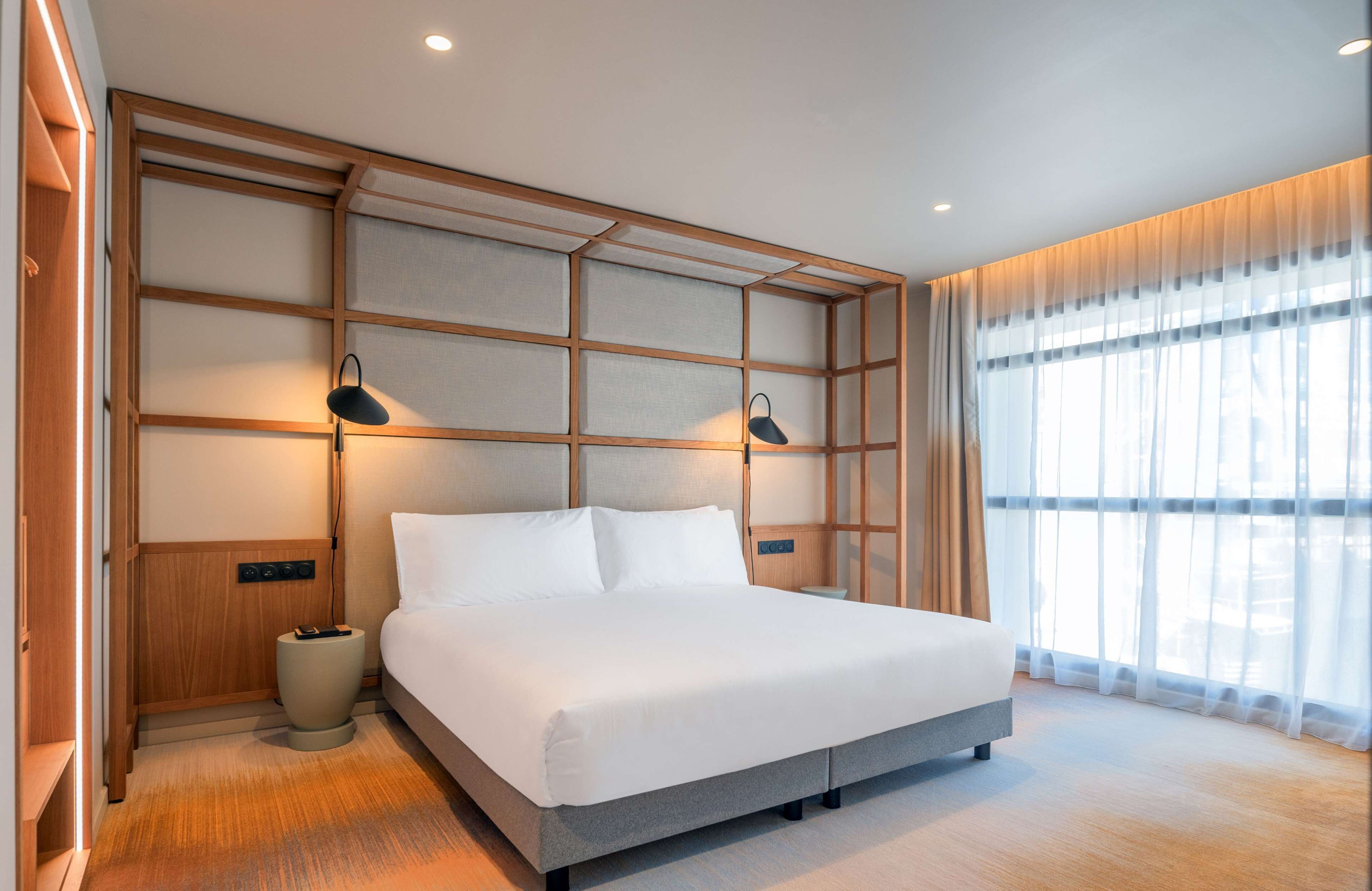 Un dormitorio moderno con una cama grande, cabecera de madera, lámparas de noche y vistas al exterior a través de cortinas translúcidas.