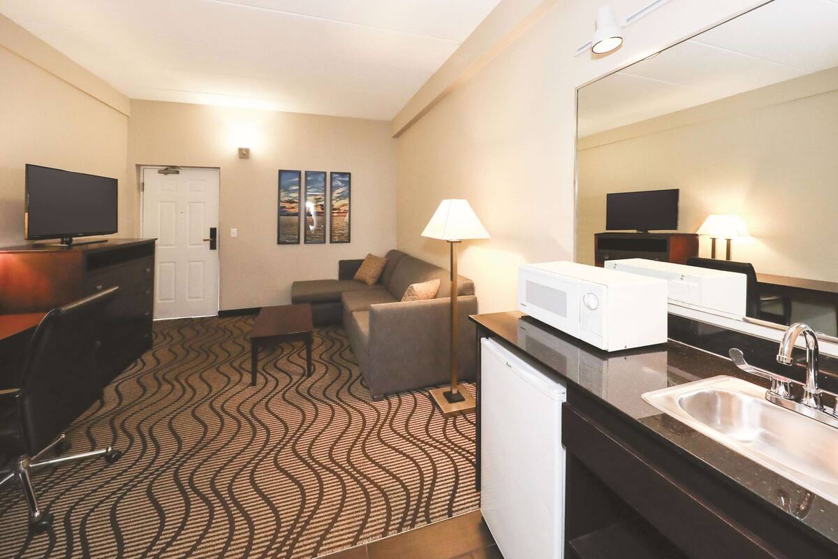 Habitación de hotel con kitchenette, sofá, televisión de pantalla plana, lámpara y un espejo grande.
