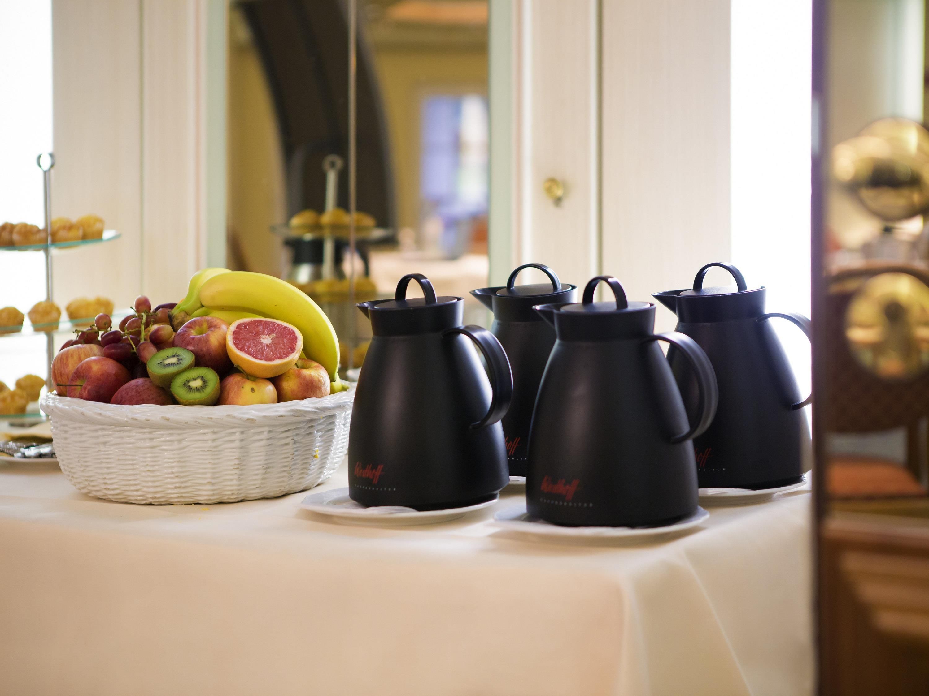 A table with a fruit basket, black coffee pots, and a clock.