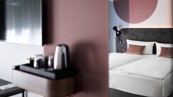 A modern hotel room with a flat-screen TV, a bedside table with a kettle, and a bed with white bedding and brown pillows.