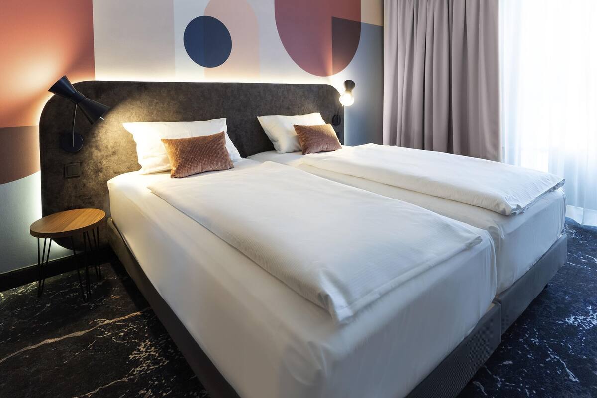 A modern hotel room with a large bed, two pillows, a bedside table, and a wall with abstract art.