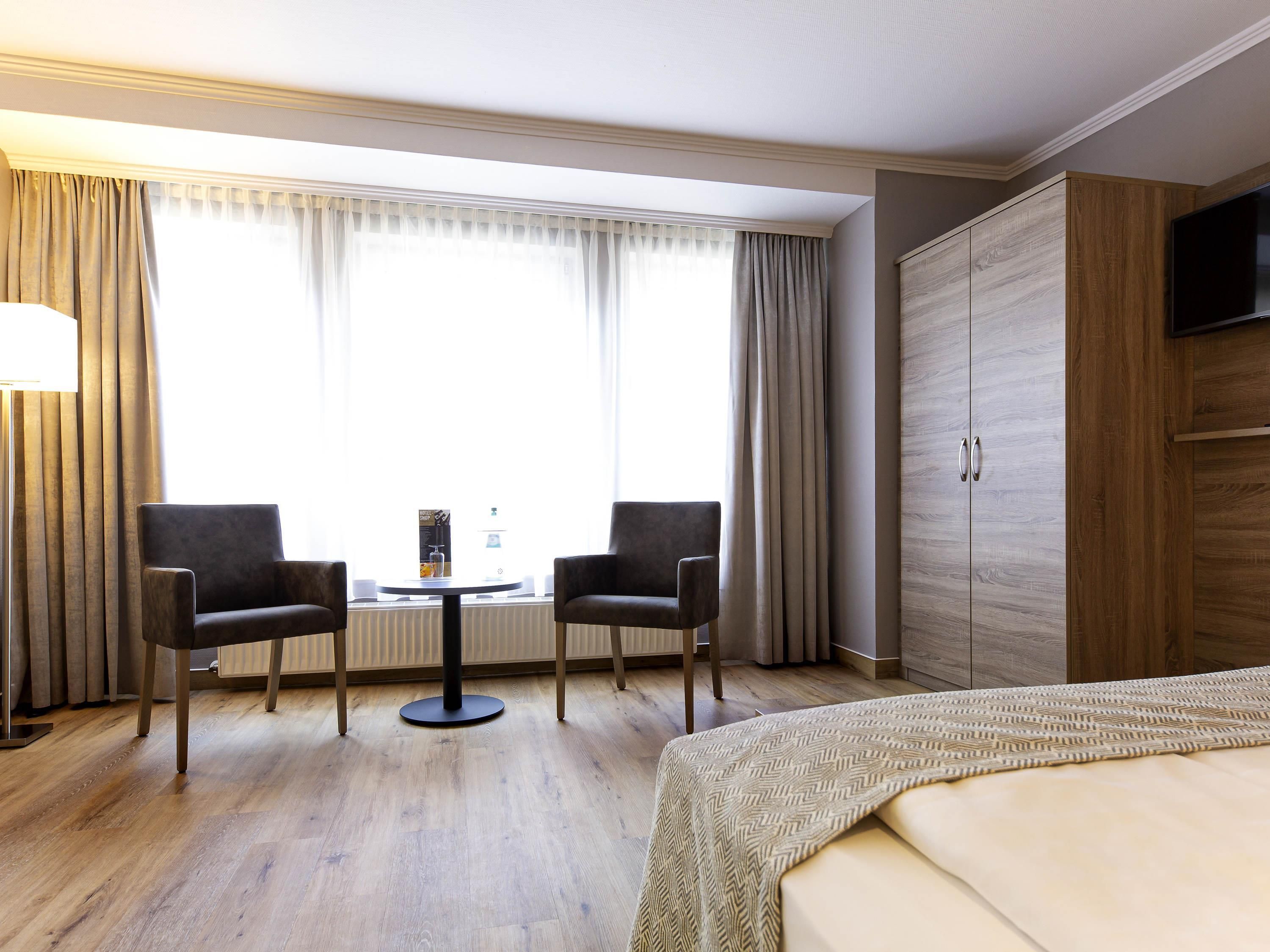 A hotel room with a bed, two armchairs, a small round table, a TV on a wooden cabinet, and a window with curtains.
