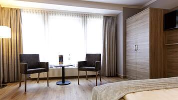 Habitación de hotel con cama, dos sillones, una mesita redonda, televisor sobre un mueble de madera y una ventana con cortinas.
