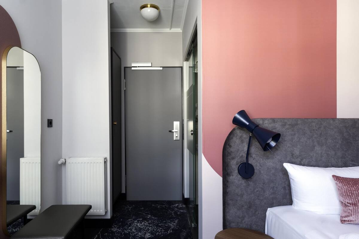 A modern hotel room with a bed, a bench, a mirror, and a wall-mounted lamp.