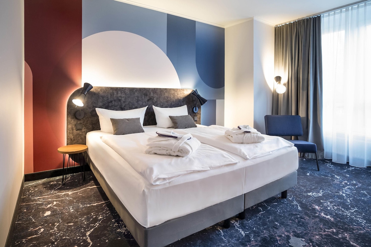 A hotel room with a large bed, a bedside table, a chair, and a view of the outdoors.