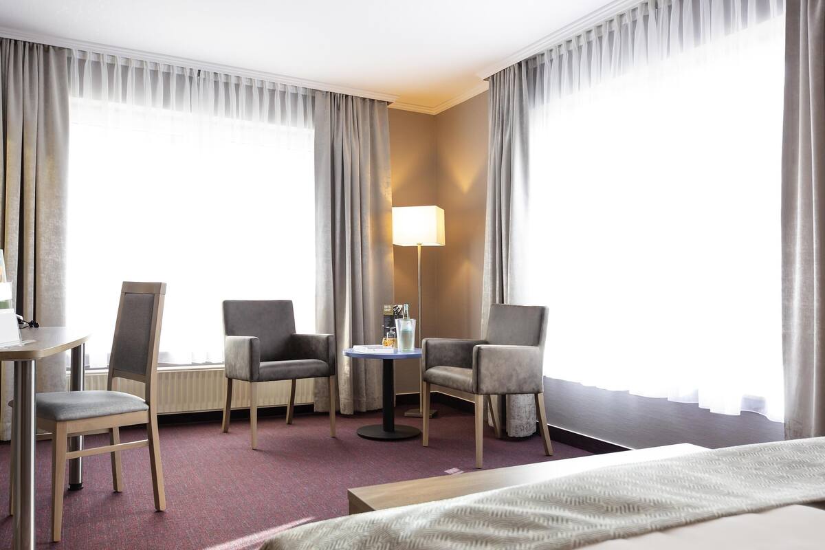 A hotel room with a bed, two armchairs, a small table, a lamp, and large windows with curtains.