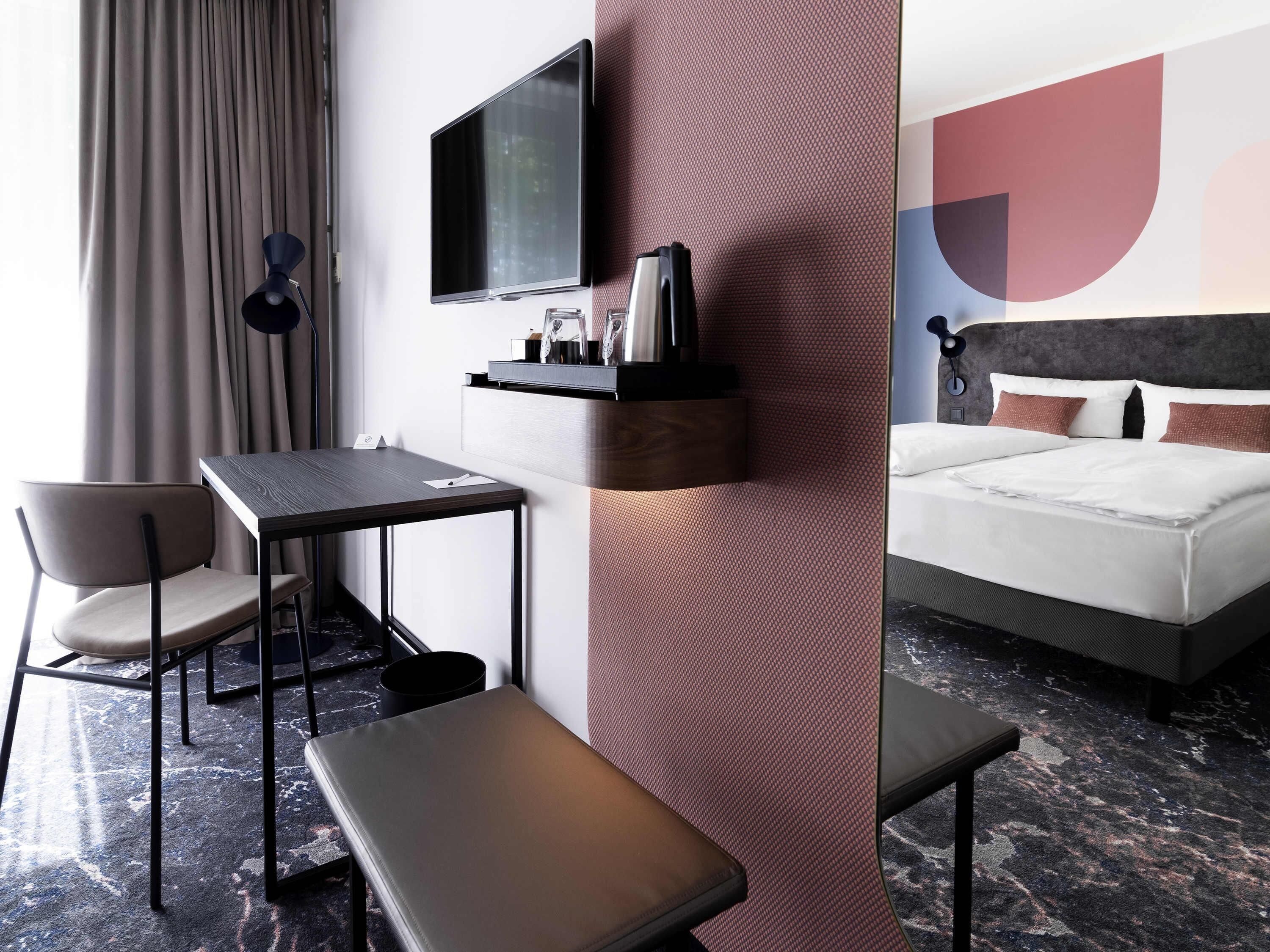 A hotel room with a bed, a desk with a chair, a bench, a TV mounted on the wall, and a large abstract wall design.