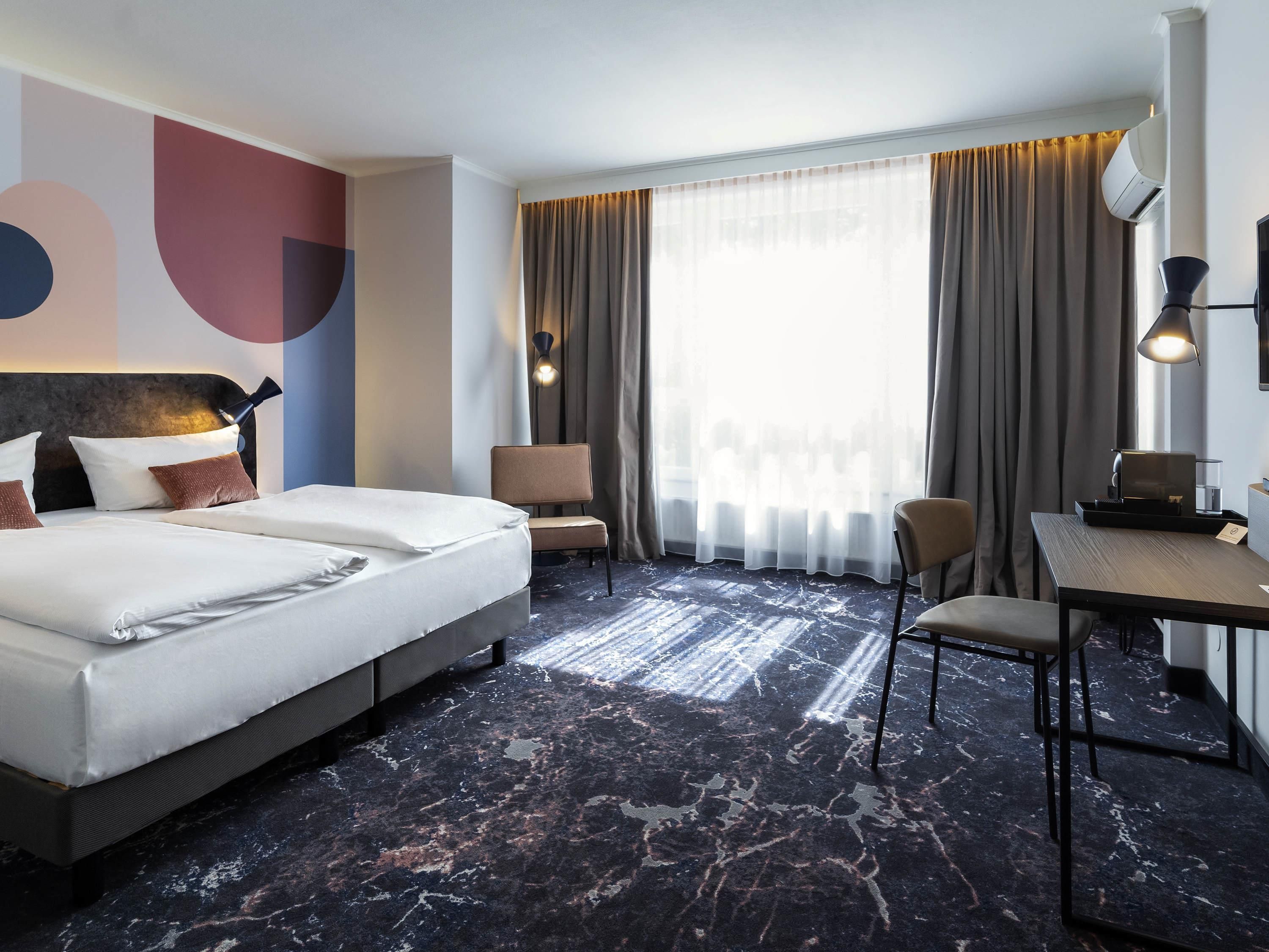A hotel room with a large bed, two chairs, a desk, and a large window with curtains.