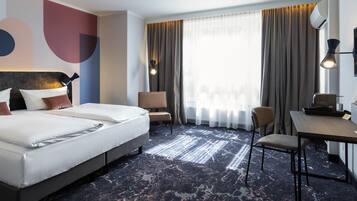 A hotel room with a large bed, two chairs, a desk, and a large window with curtains.