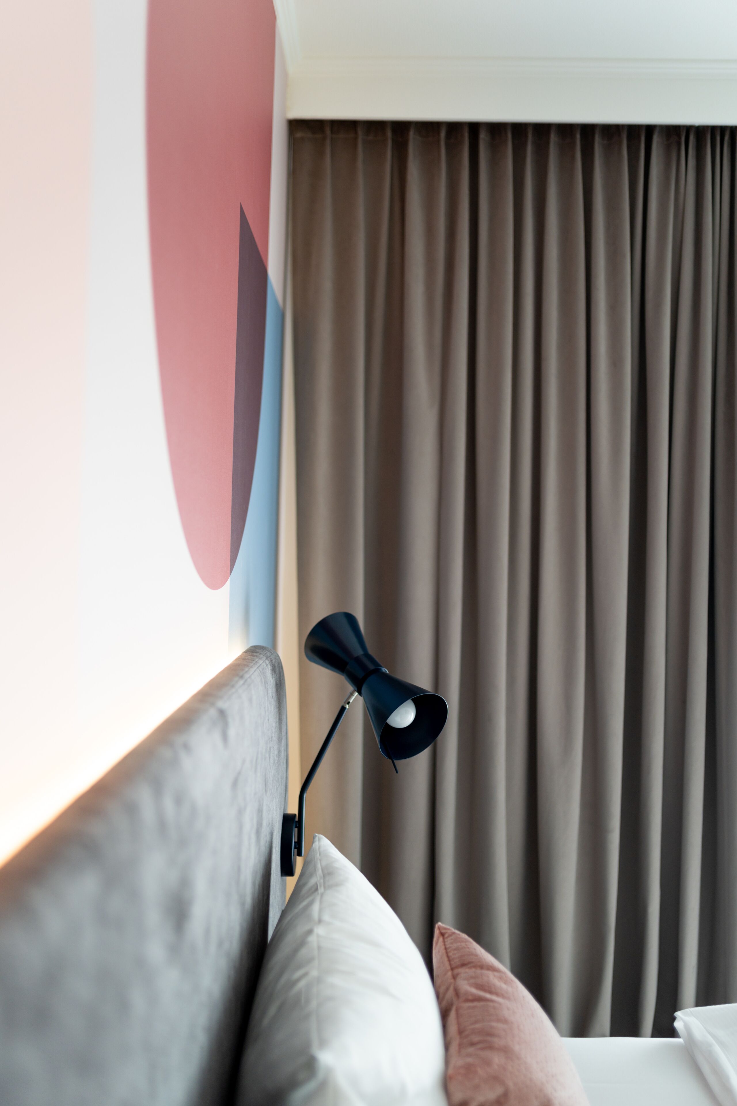 A neatly made bed with a grey headboard, a black desk lamp, and a brown curtain in the background.