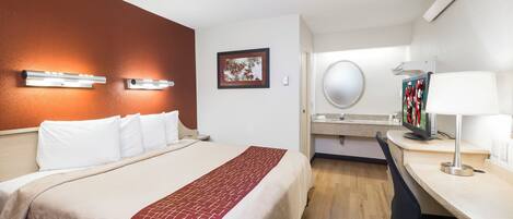 A hotel room with a bed, bedside table, desk, chair, and a bathroom with a mirror and sink.