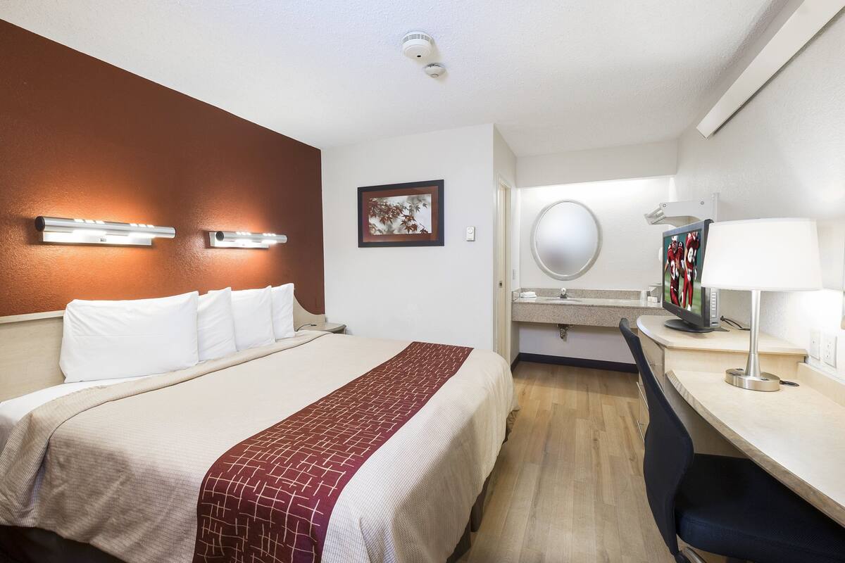 A hotel room with a bed, a desk with a chair, a mirror, and a television.