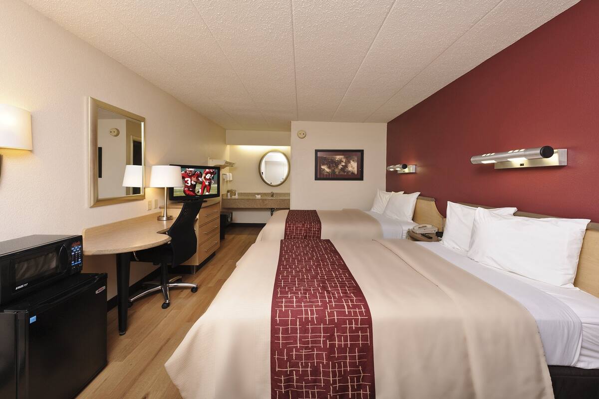 A hotel room with a bed, a desk, a chair, a microwave, a refrigerator, a mirror, a round mirror, a picture on the wall, and a wall-mounted light fixture.