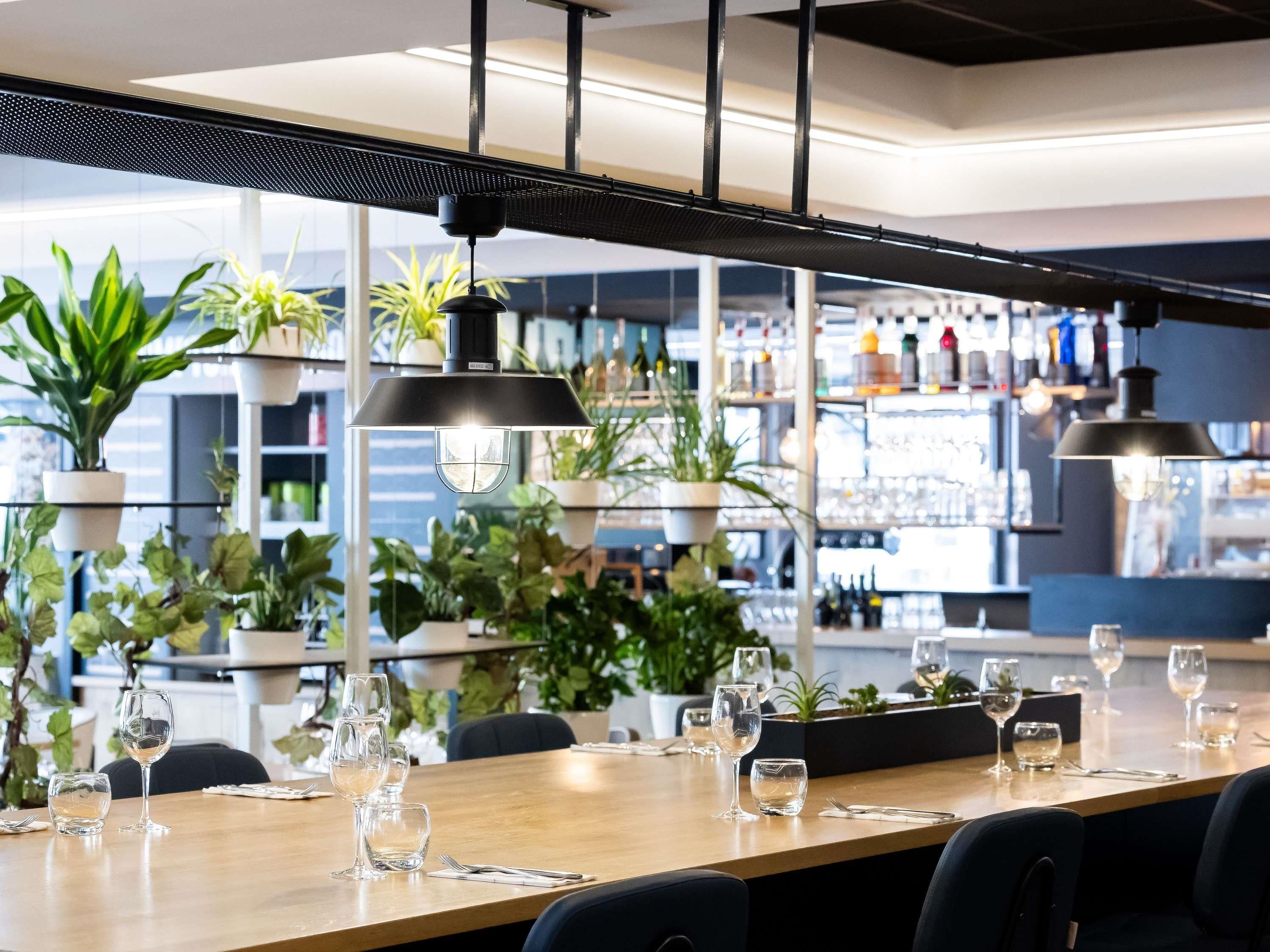 A restaurant interior with a long wooden table set for dining, surrounded by chairs, and potted plants on shelves in the background.