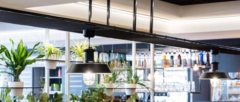 A restaurant interior with a long wooden table set for dining, surrounded by chairs, and potted plants on shelves in the background.