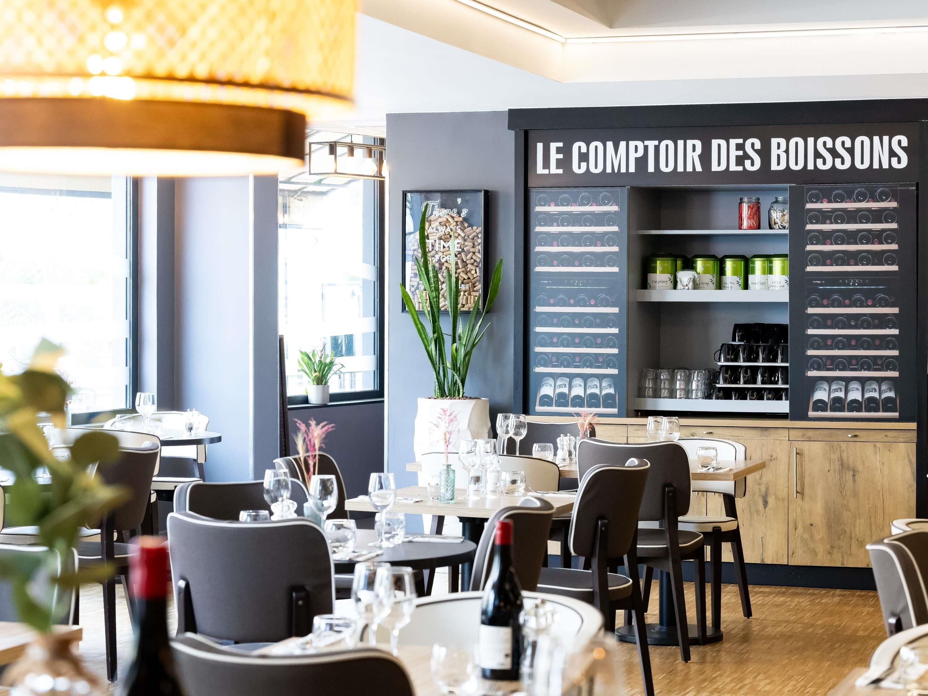 L’intérieur d’un restaurant, avec des tables dressées pour le service, un présentoir à vins et une enseigne indiquant « LE COMPTOIR DES BOISSONS ».