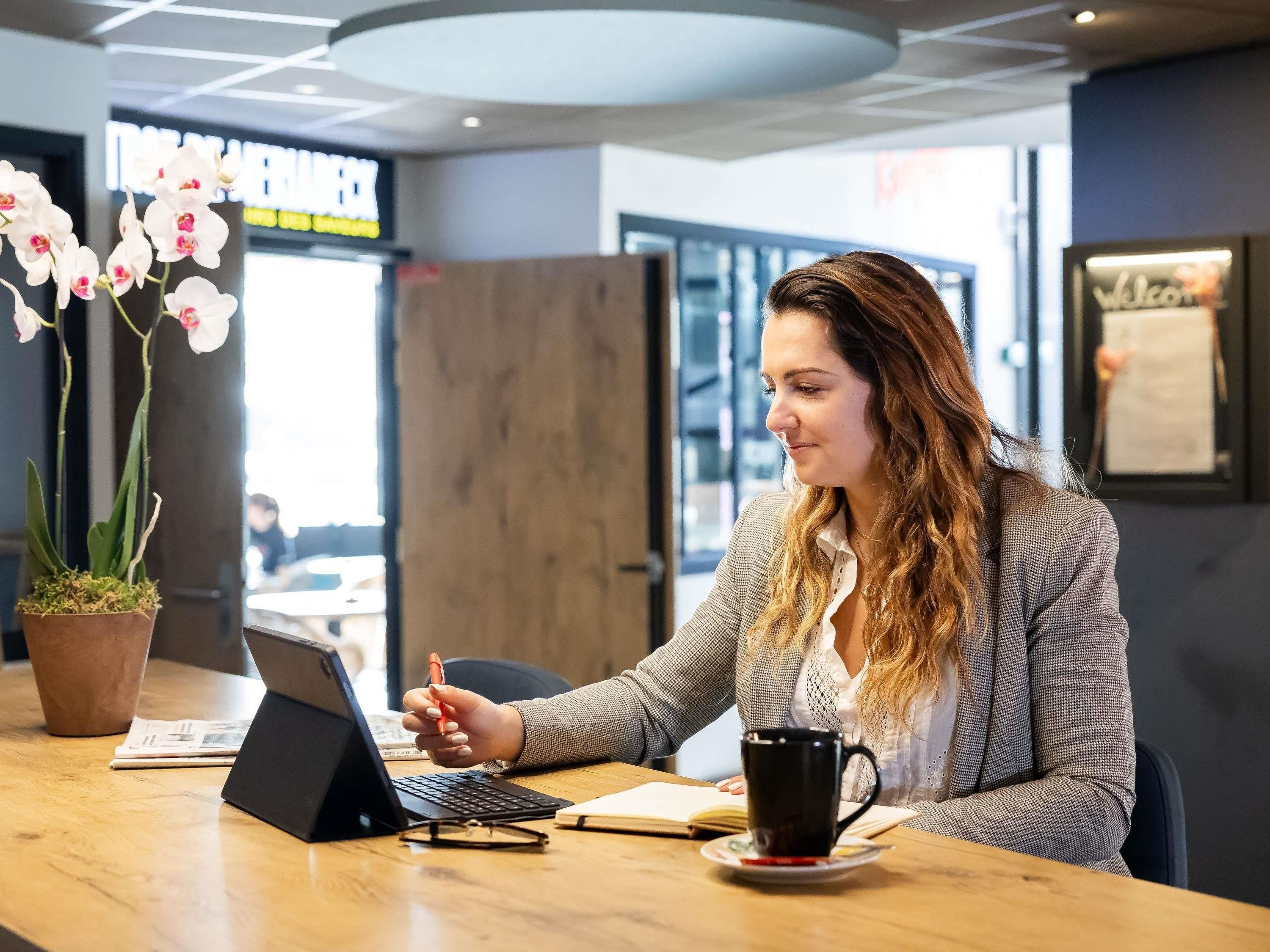 A person working at a wooden desk with a laptop, a notebook, and a coffee cup. There is a potted orchid on the left and a sign in the background.