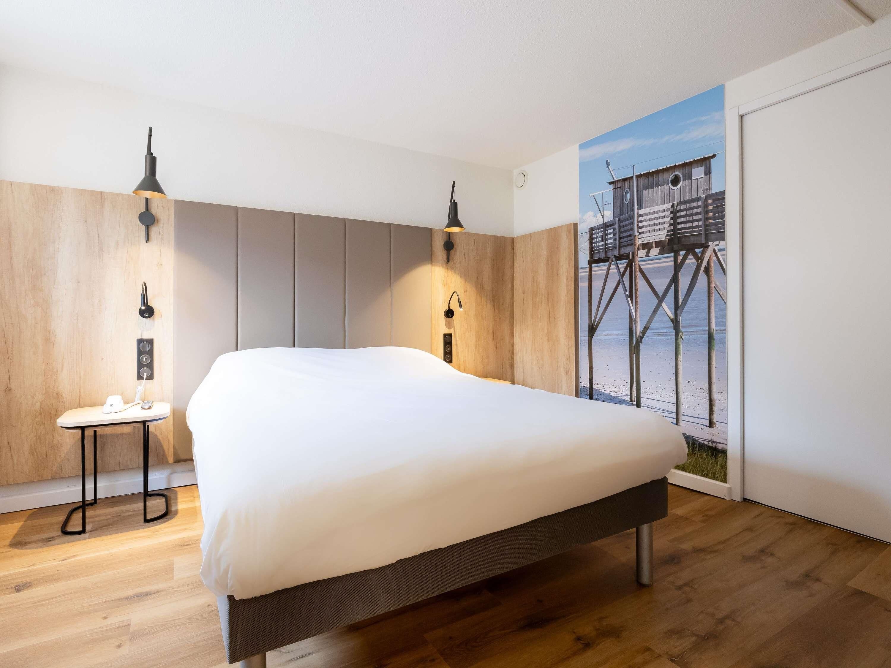 Ein Schlafzimmer mit einem Bett, zwei Nachttischen, einer Wandlampe und einem Wandbild mit Strandmotiv.