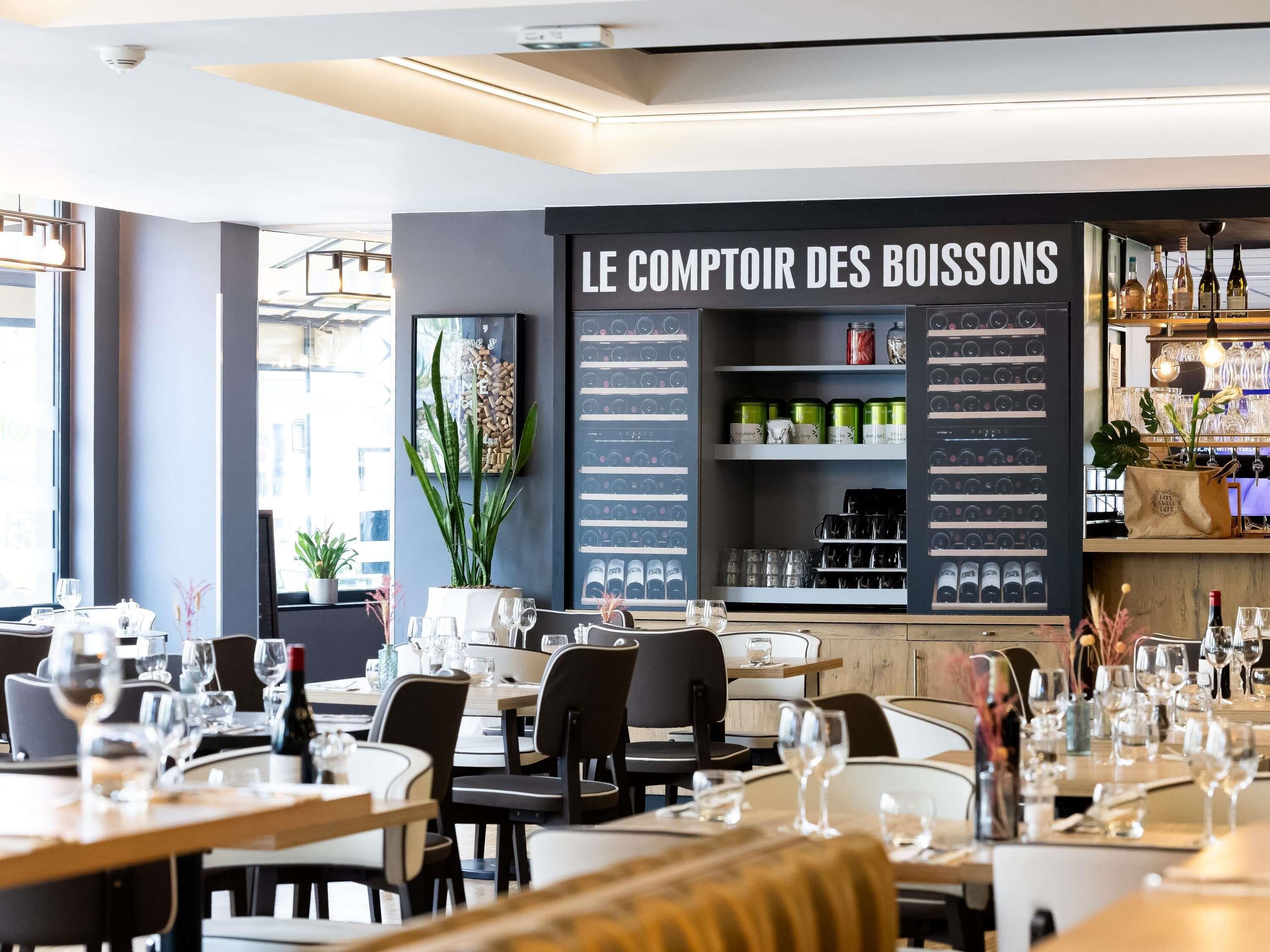 A restaurant interior with tables set for dining, a menu board titled 'LE COMPTOIR DES BOISSONS,' and shelves stocked with wine bottles and glasses.