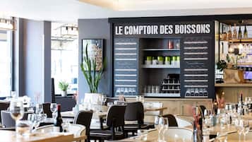 Interior de un restaurante con mesas dispuestas para comer, un cartel con el menú titulado 'LE COMPTOIR DES BOISSONS', y estantes llenos de botellas de vino y copas.