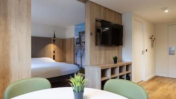 Et moderne værelse med en seng, et fjernsyn ophængt på væggen, en lille plante på en hylde og et rundt bord med stole.