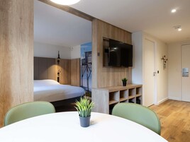 Una habitación moderna con una cama, un televisor montado en la pared, una pequeña planta en un estante y una mesa redonda con sillas.