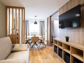 A modern living room with a sofa, a dining table with chairs, a TV mounted on the wall, and wooden shelving. - ibis Bordeaux Centre Meriadeck (Bordeaux)