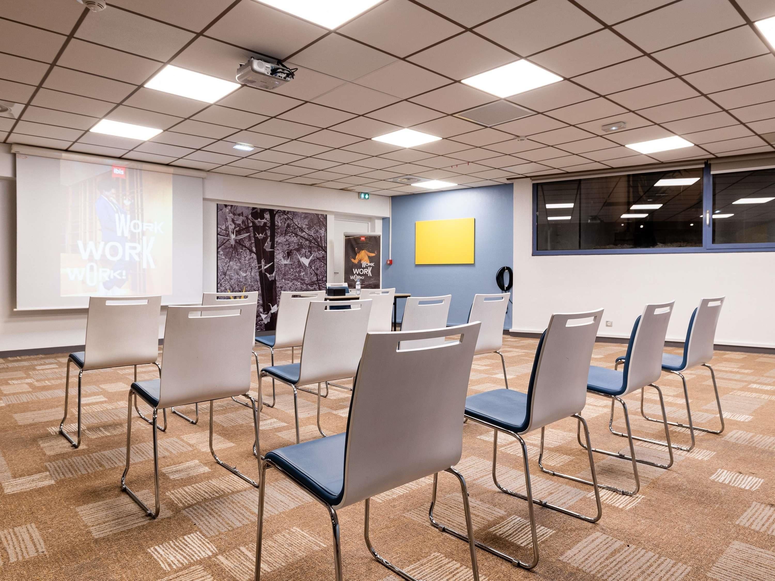 A conference room with a projector screen, white chairs, a carpeted floor, and a window.