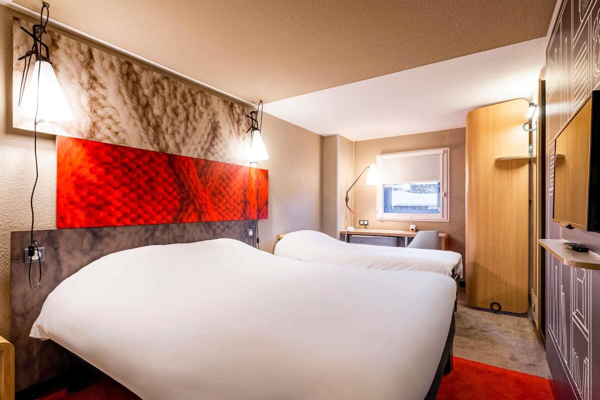 A hotel room with two beds, a desk, a window with a view, and a red carpet.