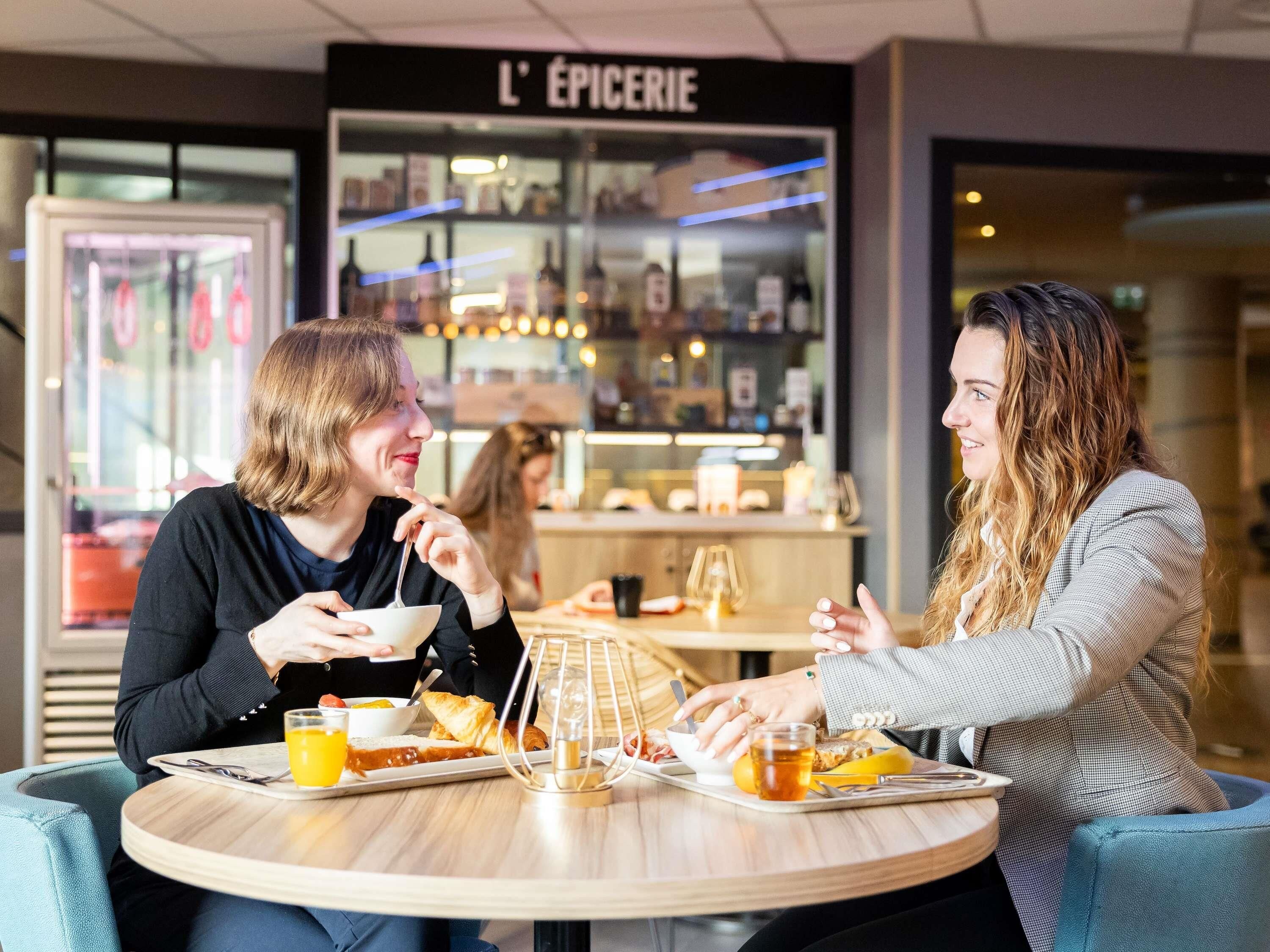 Two people dining at a table with food and drinks, a glass of orange juice, and a candle. In the background, there is a store with a sign that reads 'L'ÉPICERIE'.