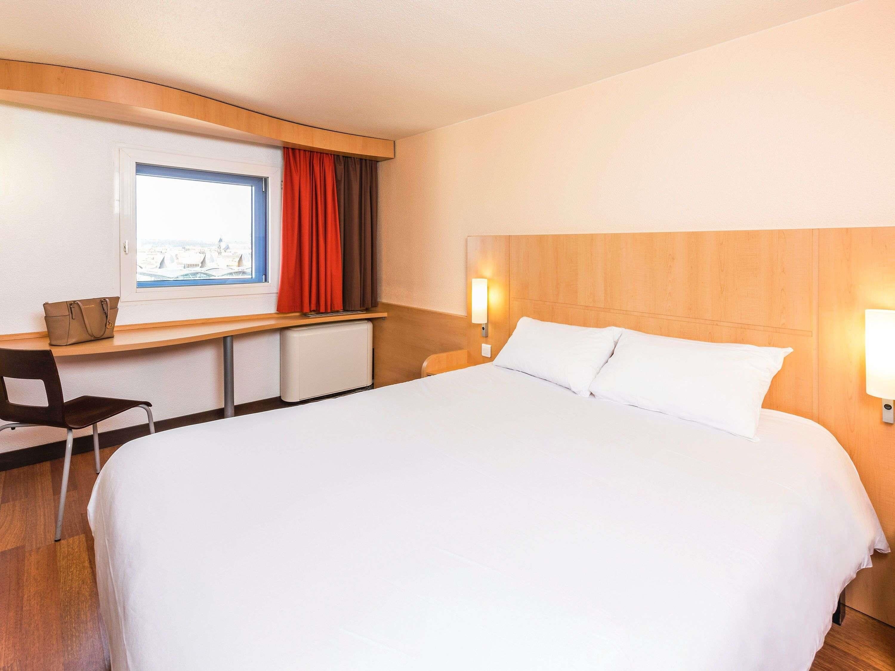 Habitación de hotel con una cama grande, un escritorio, una silla y un ventanal con cortinas.