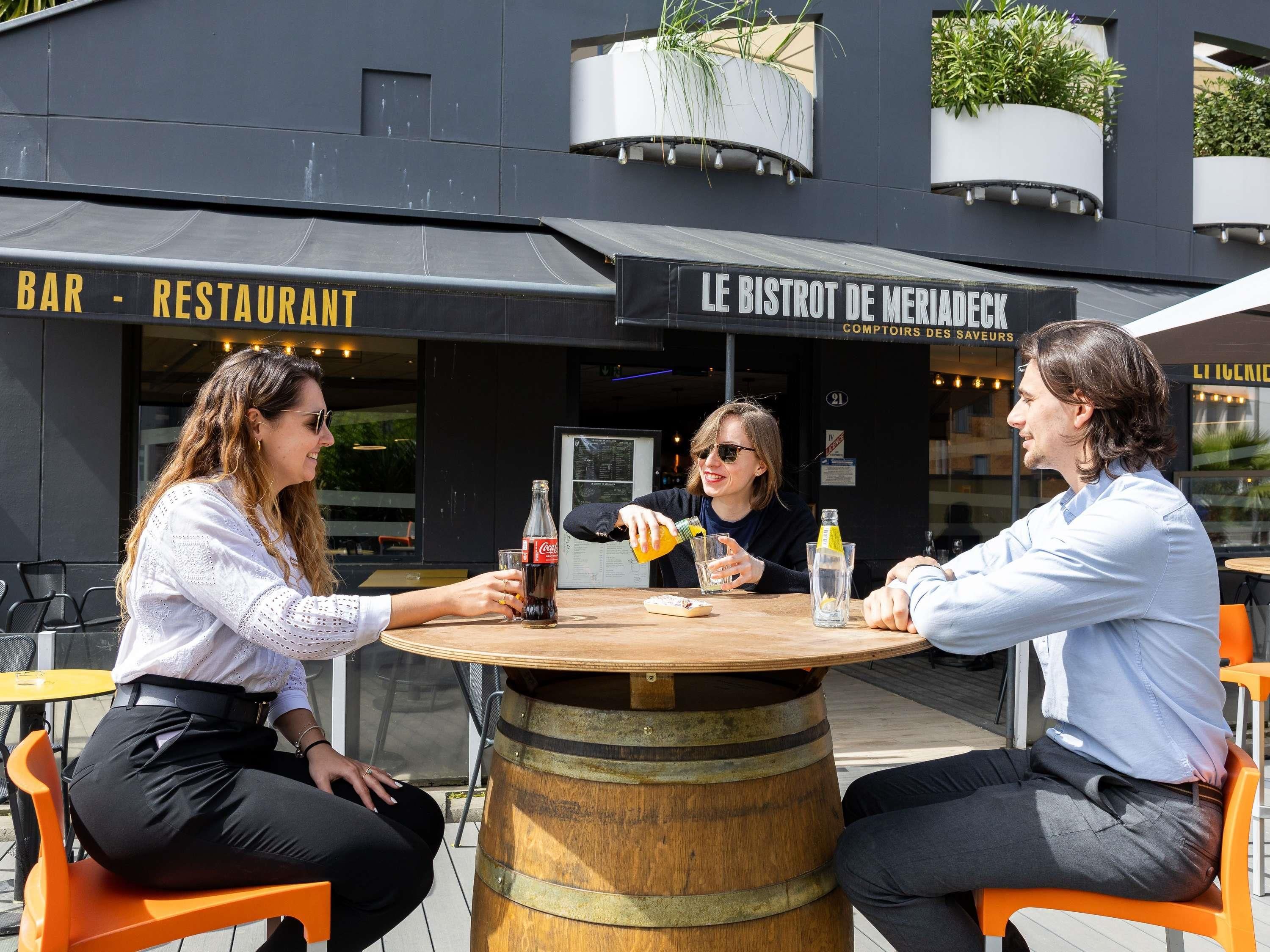 An outdoor seating area with a wooden barrel table, orange chairs, and a building with signage reading "BAR - RESTAURANT" and "LE BISTROT DE MERIADEK".